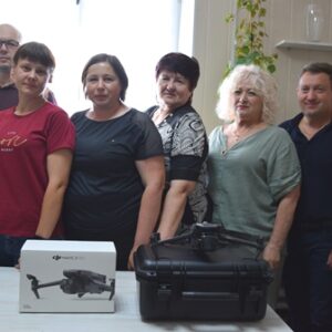 Група працівників стоїть у приміщенні за столом, на якому розміщено дрон DJI Mavic 3 Pro у коробці та кейсі для транспортування. Люди усміхаються, атмосфера дружня. Фото зроблено в офісі з яскравим природним освітленням із вікна на задньому плані