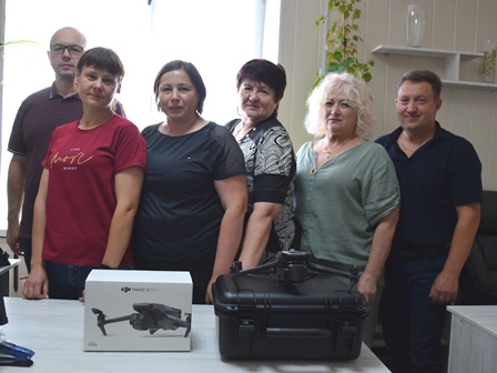 Група працівників стоїть у приміщенні за столом, на якому розміщено дрон DJI Mavic 3 Pro у коробці та кейсі для транспортування. Люди усміхаються, атмосфера дружня. Фото зроблено в офісі з яскравим природним освітленням із вікна на задньому плані