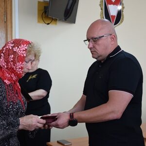 У приміщенні офіційної установи чоловік урочисто передає нагороду літній жінці в хустці. Вона тримає у руках відзнаку, ймовірно — державну. На задньому плані — ще одна жінка та емблема на стіні. Атмосфера — зворушлива та урочиста.