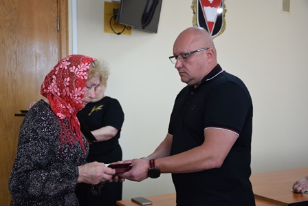 У приміщенні офіційної установи чоловік урочисто передає нагороду літній жінці в хустці. Вона тримає у руках відзнаку, ймовірно — державну. На задньому плані — ще одна жінка та емблема на стіні. Атмосфера — зворушлива та урочиста.
