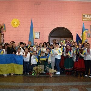 На світлині – учасники патріотичного заходу у будинку культури села Городок. Люди тримають у руках державні прапори, синьо-жовті стрічки та символіку України. На передньому плані діти з великим прапором України, позаду – жителі громади та артисти у традиційних українських костюмах. Атмосфера єдності та святкування.