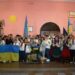На світлині – учасники патріотичного заходу у будинку культури села Городок. Люди тримають у руках державні прапори, синьо-жовті стрічки та символіку України. На передньому плані діти з великим прапором України, позаду – жителі громади та артисти у традиційних українських костюмах. Атмосфера єдності та святкування.