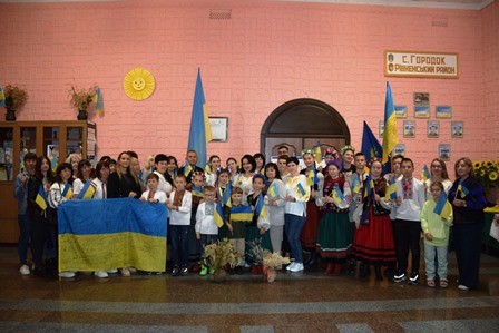На світлині – учасники патріотичного заходу у будинку культури села Городок. Люди тримають у руках державні прапори, синьо-жовті стрічки та символіку України. На передньому плані діти з великим прапором України, позаду – жителі громади та артисти у традиційних українських костюмах. Атмосфера єдності та святкування.