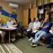 У приміщенні, прикрашеному державними прапорами України та гербом, відбувається засідання. За столом сидять чоловіки й жінка, уважно працюючи з документами. Інші учасники засідання розмістилися на стільцях уздовж стіни, деякі з них тримають папери в руках. Атмосфера зосереджена та ділова.