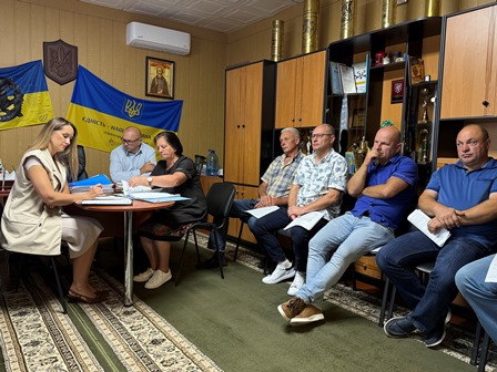 У приміщенні, прикрашеному державними прапорами України та гербом, відбувається засідання. За столом сидять чоловіки й жінка, уважно працюючи з документами. Інші учасники засідання розмістилися на стільцях уздовж стіни, деякі з них тримають папери в руках. Атмосфера зосереджена та ділова.