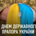 Військовослужбовець у камуфляжній формі тримає у руках жовто-блакитну тканину, на якій висить підвіска у вигляді тризуба — герба України. Внизу зображення напис: «З Днем Державного Прапора України».