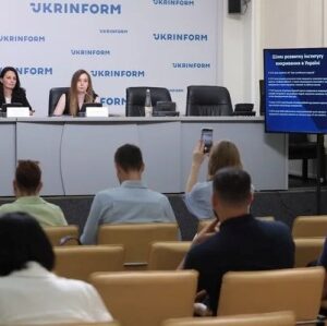 Пресконференція в інформаційному агентстві «Укрінформ». Двоє жінок сидять за столом на сцені перед мікрофонами, поруч екран із презентацією. У залі кілька слухачів, один із них фотографує захід на телефон. На стіні позаду розміщено логотип «Укрінформ»