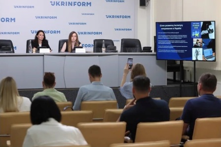 Пресконференція в інформаційному агентстві «Укрінформ». Двоє жінок сидять за столом на сцені перед мікрофонами, поруч екран із презентацією. У залі кілька слухачів, один із них фотографує захід на телефон. На стіні позаду розміщено логотип «Укрінформ»