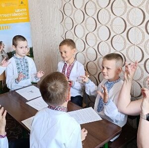 Група дітей у вишиванках бере участь у занятті в Центрі життєстійкості Городоцької територіальної громади. Діти сидять за столом разом із педагогом і виконують вправи руками. На стіні — інформаційний банер Центру.