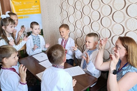 Група дітей у вишиванках бере участь у занятті в Центрі життєстійкості Городоцької територіальної громади. Діти сидять за столом разом із педагогом і виконують вправи руками. На стіні — інформаційний банер Центру.