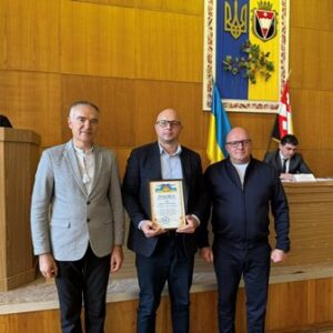 У залі засідань із дерев’яними панелями на стіні троє чоловіків стоять перед трибуною. Один із них тримає у руках грамоту. Позаду видно прапори України та Рівненщини, а також герб і державний прапор.