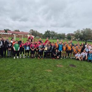 Велика група дітей та підлітків у спортивній формі стоїть на футбольному полі після змагань. Учасники поділені на команди в різнокольоровій формі (чорній, рожевій, помаранчевій, білій та іншій). Багато з них тримають медалі та грамоти.