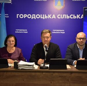 На фото троє представників Городоцької сільської ради сидять за столом під час офіційного засідання. На стіні позаду — синій банер із написом «Городоцька сільська рада» та гербом. Ліворуч встановлено прапор України.