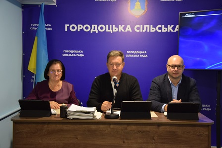 На фото троє представників Городоцької сільської ради сидять за столом під час офіційного засідання. На стіні позаду — синій банер із написом «Городоцька сільська рада» та гербом. Ліворуч встановлено прапор України.