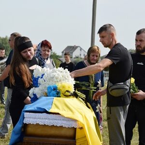 Люди прощаються із загиблим воїном під час похорону. На передньому плані труна, накрита Державним Прапором України та квітами. Рідні та побратими кладуть квіти, висловлюючи шану та скорботу.