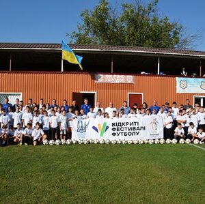 Велика група дітей та дорослих у білих футболках стоять на футбольному полі перед будівлею стадіону. Попереду розтягнутий банер із написом «Відкритий фестиваль футболу» та логотипами. На даху будівлі майорить державний прапор України. На задньому плані видно вивіску «ФК Городок»