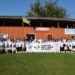 Велика група дітей та дорослих у білих футболках стоять на футбольному полі перед будівлею стадіону. Попереду розтягнутий банер із написом «Відкритий фестиваль футболу» та логотипами. На даху будівлі майорить державний прапор України. На задньому плані видно вивіску «ФК Городок»