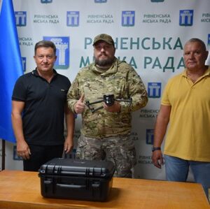 Троє чоловіків стоять у приміщенні на фоні банера з написом «Рівненська міська рада». Посередині – військовослужбовець у камуфляжній формі з дронами у руках, поруч із ним двоє чоловіків у цивільному одязі. На столі перед ними чорний кейс. Ліворуч і праворуч встановлені прапори.