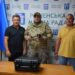 Троє чоловіків стоять у приміщенні на фоні банера з написом «Рівненська міська рада». Посередині – військовослужбовець у камуфляжній формі з дронами у руках, поруч із ним двоє чоловіків у цивільному одязі. На столі перед ними чорний кейс. Ліворуч і праворуч встановлені прапори.