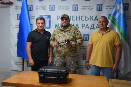 Троє чоловіків стоять у приміщенні на фоні банера з написом «Рівненська міська рада». Посередині – військовослужбовець у камуфляжній формі з дронами у руках, поруч із ним двоє чоловіків у цивільному одязі. На столі перед ними чорний кейс. Ліворуч і праворуч встановлені прапори.