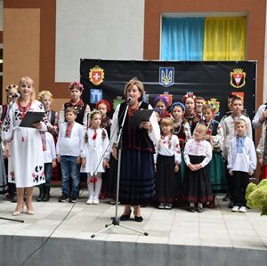 На сцені стоїть група дітей та кілька дорослих у традиційному українському вбранні — вишиванках, спідницях, камізельках та віночках із різнокольоровими стрічками. Двоє жінок тримають у руках мікрофони й читають або співають. Позаду розміщений чорний банер із гербами міст та символікою України: державний герб, прапор та синьо-жовті кольори.