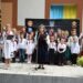 На сцені стоїть група дітей та кілька дорослих у традиційному українському вбранні — вишиванках, спідницях, камізельках та віночках із різнокольоровими стрічками. Двоє жінок тримають у руках мікрофони й читають або співають. Позаду розміщений чорний банер із гербами міст та символікою України: державний герб, прапор та синьо-жовті кольори.