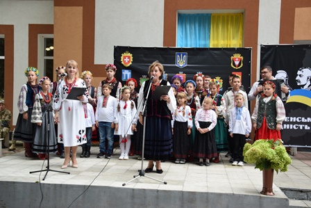 На сцені стоїть група дітей та кілька дорослих у традиційному українському вбранні — вишиванках, спідницях, камізельках та віночках із різнокольоровими стрічками. Двоє жінок тримають у руках мікрофони й читають або співають. Позаду розміщений чорний банер із гербами міст та символікою України: державний герб, прапор та синьо-жовті кольори.