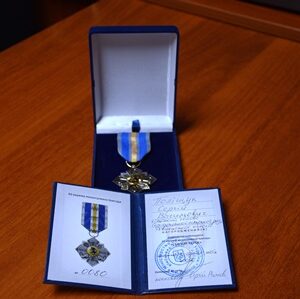 На дерев’яному столі розташована відзнака у футлярі синього кольору. Усередині лежить медаль із синьо-жовтою стрічкою та зображенням емблеми. Поруч відкрите посвідчення з печаткою та підписом, де зазначено ім’я нагородженого.