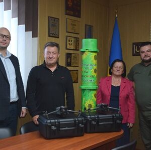 У кабінеті біля столу стоїть четверо людей: двоє чоловіків у цивільному одязі, жінка в рожевому піджаку та чоловік у військовій формі. Перед ними на столі розташовані дві чорні валізи з технікою