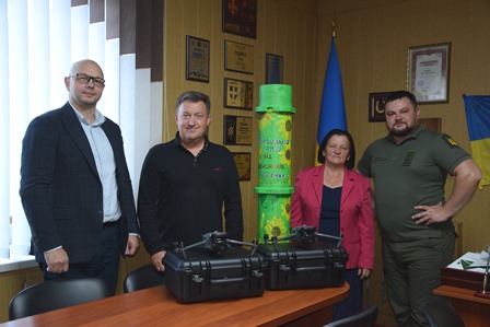 У кабінеті біля столу стоїть четверо людей: двоє чоловіків у цивільному одязі, жінка в рожевому піджаку та чоловік у військовій формі. Перед ними на столі розташовані дві чорні валізи з технікою