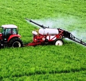 Червоний трактор із причіпним обприскувачем працює на зеленому полі, розпилюючи засіб захисту рослин.