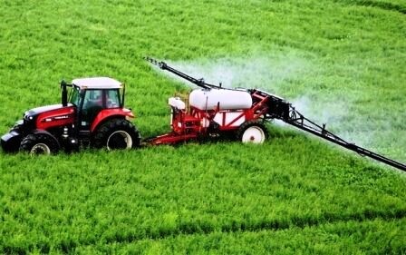 Червоний трактор із причіпним обприскувачем працює на зеленому полі, розпилюючи засіб захисту рослин.