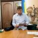 Чоловік у світлій сорочці сидить за столом та читає документи під час засідання. На столі лежать папери й фотокамера, позаду видно український прапор та елементи декору