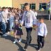 Учень-випускник у вишиванці з першокласниками веде їх урочистою ходою на святі першого дзвоника. Дівчинка тримає дзвоник, поруч йде хлопчик. На задньому плані – учні та батьки.