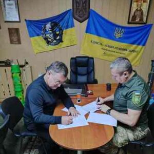 На фото двоє чоловіків сидять за столом і підписують документи. На стіні за ними розміщено два українські прапори з написами та герб України. Один із чоловіків у військовій формі, інший — у цивільному одязі. Сцена відбувається в службовому кабінеті.