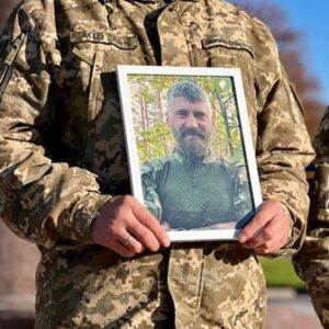 На зображенні видно військового у камуфляжній формі, який тримає в руках фотографію чоловіка у зеленому светрі на тлі дерев. Поруч стоїть ще один військовослужбовець у схожій формі. Фото зроблене на вулиці, ймовірно, під час вшанування пам’яті полеглого воїна. На тлі — ясне небо та осінні дерева.