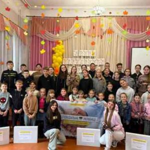На фото група дітей та підлітків у святково прикрашеній залі. На задньому плані — кольорова завіса, гірлянди з осіннім листям і жовті кульки. У центрі діти тримають банер із написом «Разом до перемоги України!». Перед ними стоять великі коробки з написами — ймовірно, це подарунки або гуманітарна допомога.