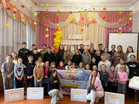 На фото група дітей та підлітків у святково прикрашеній залі. На задньому плані — кольорова завіса, гірлянди з осіннім листям і жовті кульки. У центрі діти тримають банер із написом «Разом до перемоги України!». Перед ними стоять великі коробки з написами — ймовірно, це подарунки або гуманітарна допомога.