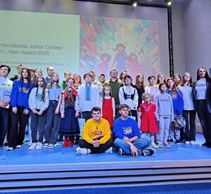 Група дітей та дорослих стоїть на сцені перед екраном із написом International Junior Contest #U_Hero Award 2026. Учасники різного віку позують для спільного фото, деякі сидять попереду, решта стоїть. На екрані позаду видно ілюстрацію з яскравими кольорами. Атмосфера святкова та урочиста.