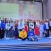 Група дітей та дорослих стоїть на сцені перед екраном із написом International Junior Contest #U_Hero Award 2026. Учасники різного віку позують для спільного фото, деякі сидять попереду, решта стоїть. На екрані позаду видно ілюстрацію з яскравими кольорами. Атмосфера святкова та урочиста.