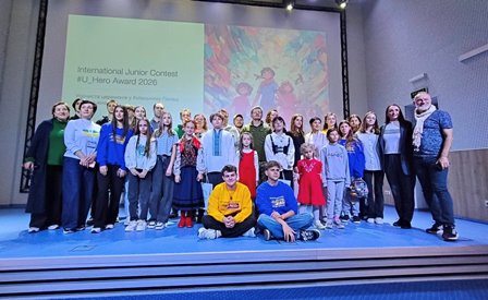 Група дітей та дорослих стоїть на сцені перед екраном із написом International Junior Contest #U_Hero Award 2026. Учасники різного віку позують для спільного фото, деякі сидять попереду, решта стоїть. На екрані позаду видно ілюстрацію з яскравими кольорами. Атмосфера святкова та урочиста.