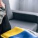 Жінка тримає в руках військову кепку в камуфляжному забарвленні, притискаючи її до обличчя. Поруч на дивані лежить складений прапор України та військова форма. Атмосфера фото передає сум і тривогу за військових