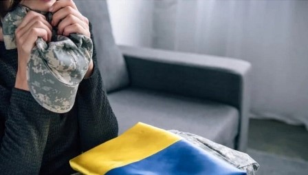 Жінка тримає в руках військову кепку в камуфляжному забарвленні, притискаючи її до обличчя. Поруч на дивані лежить складений прапор України та військова форма. Атмосфера фото передає сум і тривогу за військових