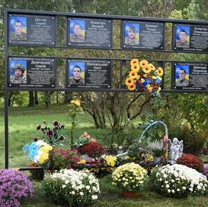 Меморіальний стенд із фотографіями та іменами загиблих українських воїнів, прикрашений живими квітами та вінками. Пам’ятне місце вшанування героїв, розташоване серед дерев та зелені."