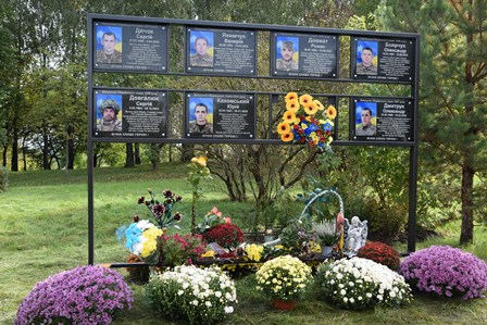 Меморіальний стенд із фотографіями та іменами загиблих українських воїнів, прикрашений живими квітами та вінками. Пам’ятне місце вшанування героїв, розташоване серед дерев та зелені."