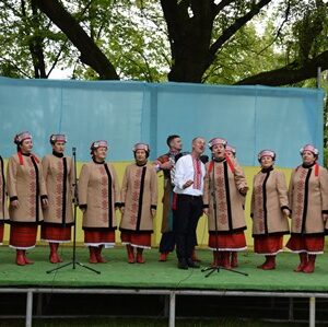 На фото — фольклорний колектив у традиційному українському вбранні виступає на сцені просто неба. Чоловік і жінки стоять у ряд, співаючи пісню.