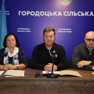 На зображенні троє представників Городоцької сільської ради сидять за столом під час засідання. Посередині — сільський голова, поруч із ним – працівники ради. На стіні позаду розміщено синє тло з написом «Городоцька сільська рада» та державним прапором України.