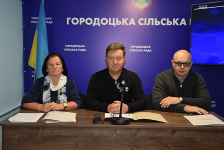 На зображенні троє представників Городоцької сільської ради сидять за столом під час засідання. Посередині — сільський голова, поруч із ним – працівники ради. На стіні позаду розміщено синє тло з написом «Городоцька сільська рада» та державним прапором України.