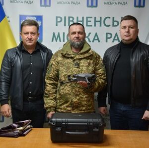 На фото троє чоловіків стоять біля столу, на якому розміщено дрон і кейс до нього. На задньому плані — прапори України та Рівненської міської ради, а також банер із написом «Рівненська міська рада»