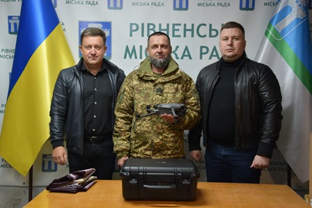 На фото троє чоловіків стоять біля столу, на якому розміщено дрон і кейс до нього. На задньому плані — прапори України та Рівненської міської ради, а також банер із написом «Рівненська міська рада»